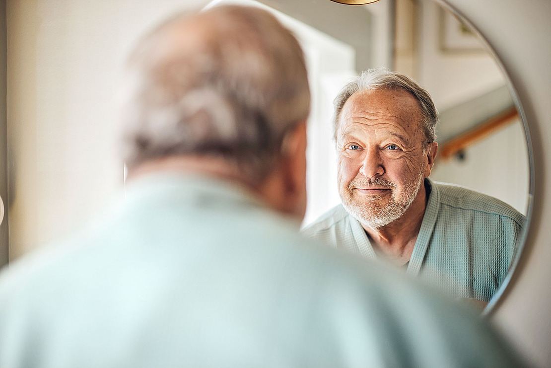 Ein älterer Mann mit grauem Haar und Bart lächelt sanft, während er sich in einem runden Spiegel betrachtet. Er trägt einen hellgrünen Bademantel und steht in einem sanft beleuchteten Raum.