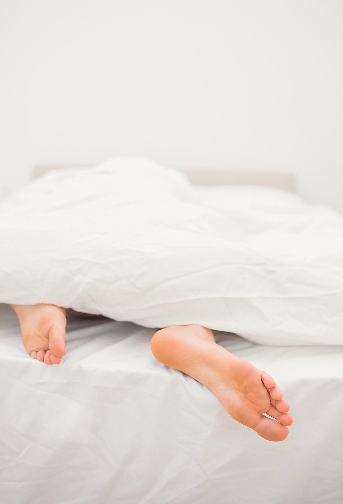 Zwei nackte Füße ragen unter einer weißen Decke auf einem ordentlich gemachten Bett hervor, das einen hellen, minimal weißen Hintergrund hat.