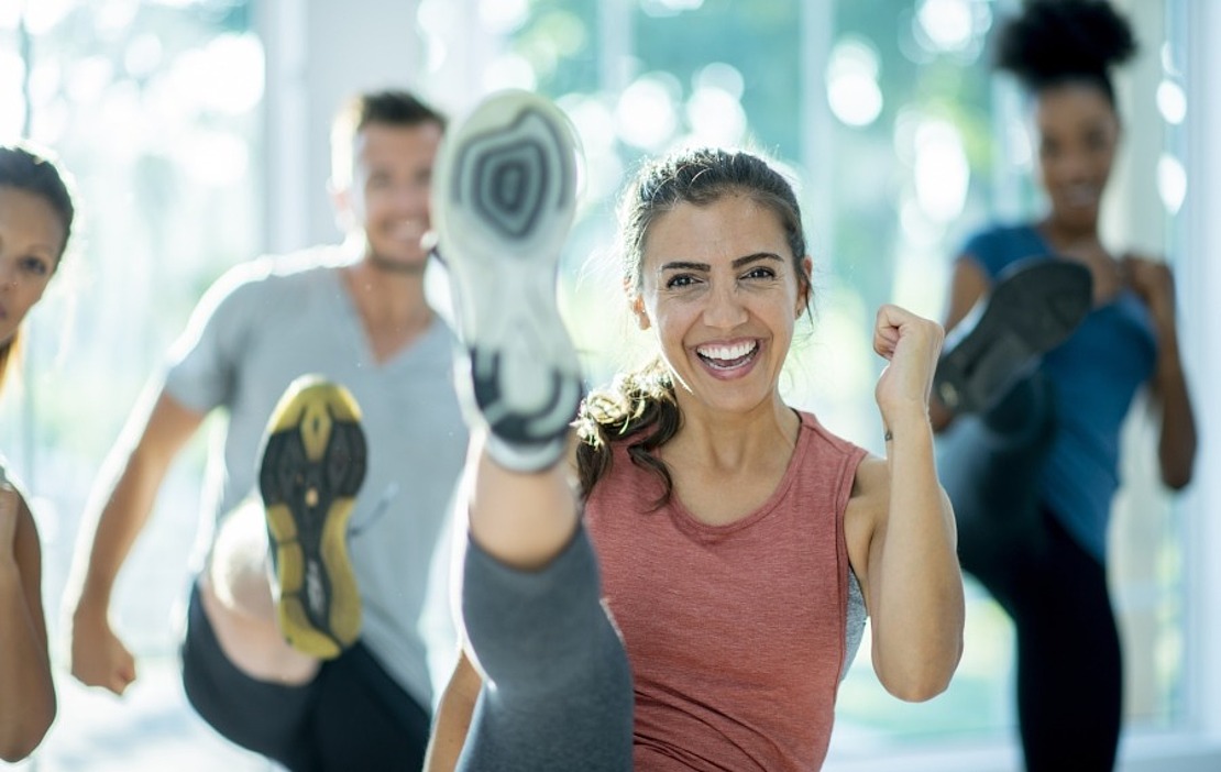 Vier Personen in Sportkleidung trainieren in einer Halle, wobei sie energisch die Beine hochwerfen und lächeln, während im Hintergrund ein sonniges Fenster zu sehen ist. Die Szene vermittelt Energie, Fitness und Begeisterung für das Gruppentraining.