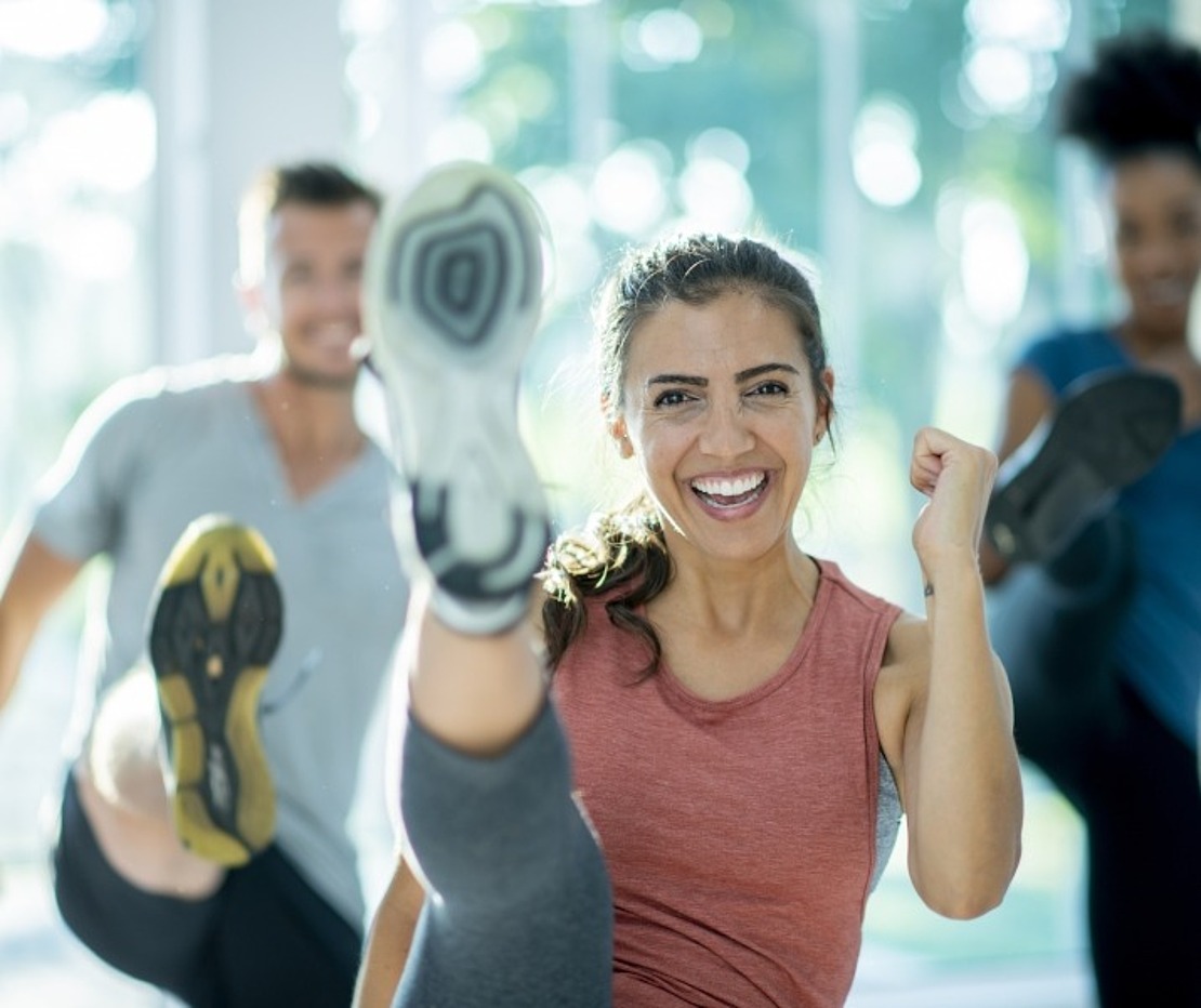 Vier Personen in Sportkleidung trainieren in einer Halle, wobei sie energisch die Beine hochwerfen und lächeln, während im Hintergrund ein sonniges Fenster zu sehen ist. Die Szene vermittelt Energie, Fitness und Begeisterung für das Gruppentraining.
