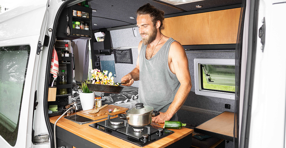 Ein Mann mit Bart und zurückgebundenem Haar kocht in einer modernen Wohnmobilküche und hält eine Pfanne mit Essen über den Herd. Der kompakte Raum ist mit Regalen, einem Fenster und Küchenutensilien ausgestattet und bietet einen gemütlichen, organisierten Kochbereich.