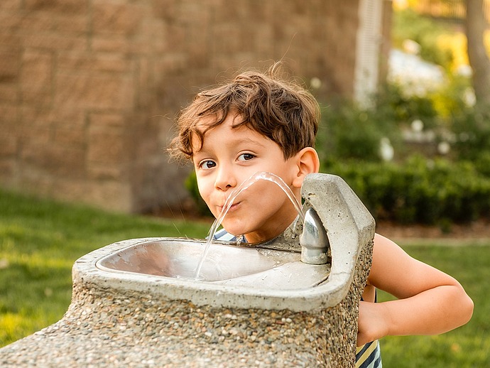 Ein kleines Kind mit lockigem braunem Haar trinkt im Freien Wasser aus einem steinernen Trinkbrunnen, im Hintergrund sind grünes Gras und eine Backsteinmauer zu sehen.
