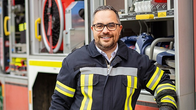 Ein Feuerwehrmann in einer dunklen Uniform mit gelben Reflexionsstreifen steht lächelnd neben einem offenen Feuerwehrauto, das mit Rettungsgeräten gefüllt ist.