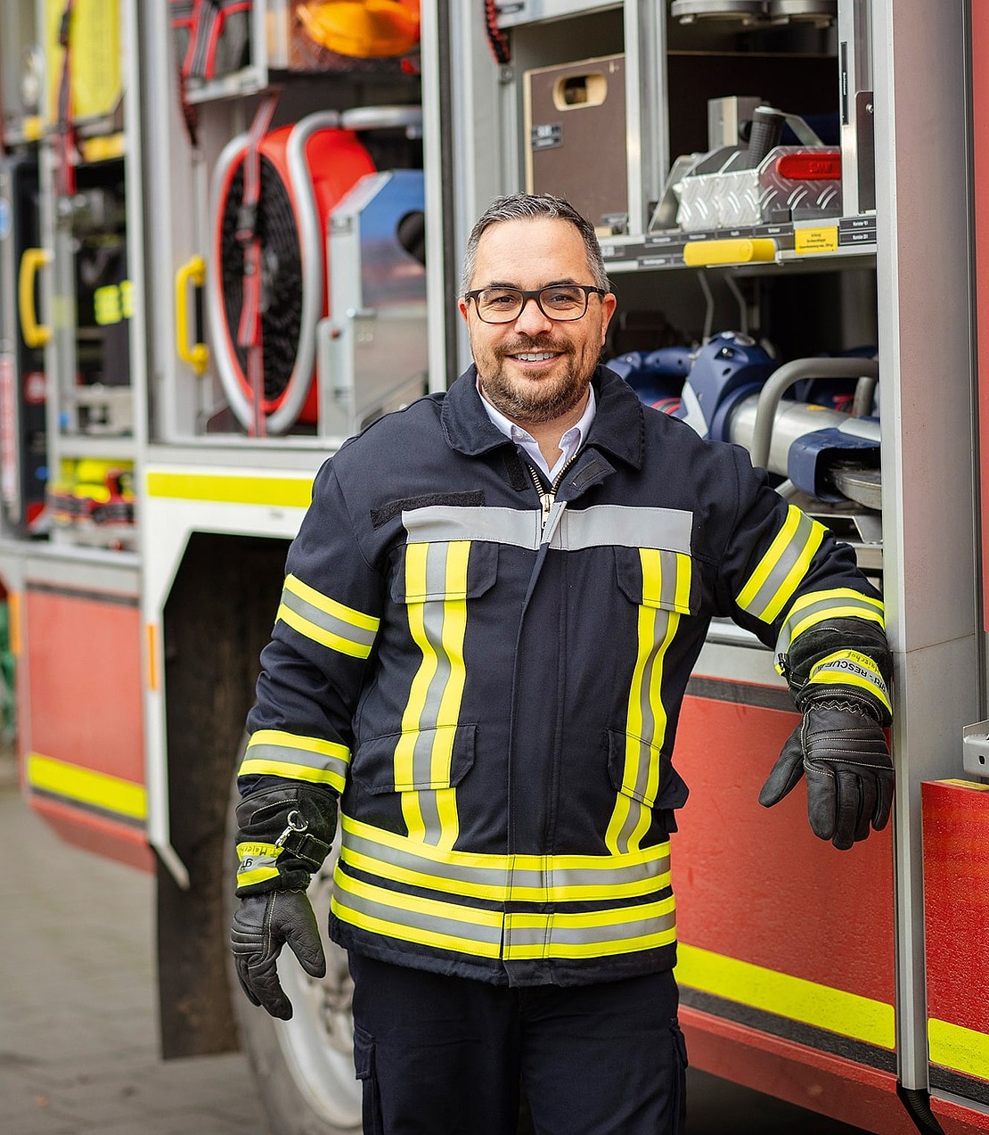 Ein Feuerwehrmann in einer dunklen Uniform mit gelben Reflexionsstreifen steht lächelnd neben einem offenen Feuerwehrauto, das mit Rettungsgeräten gefüllt ist.
