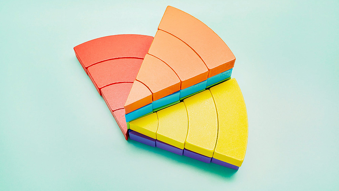 Bunte Halbkreisblöcke aus Holz in Rot-, Orange- und Gelbtönen sind fächerförmig auf einem hellblauen Hintergrund angeordnet. Die Teile passen wie Puzzle-Scheiben zusammen und bilden ein geometrisches Muster.