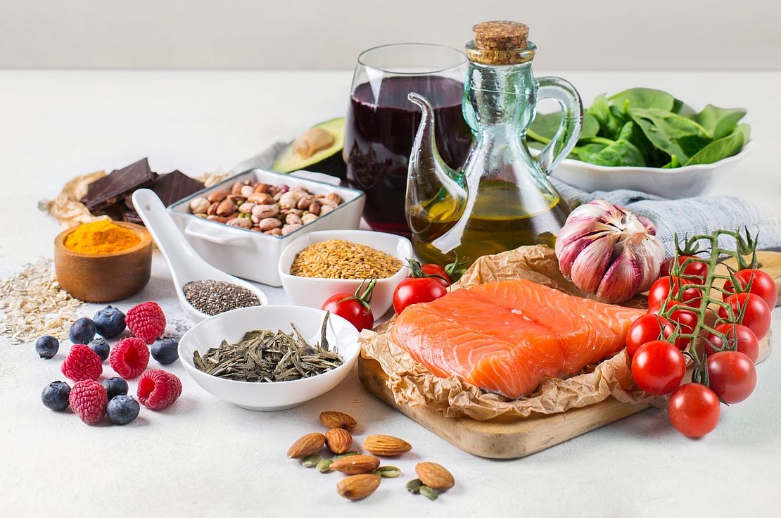 Eine Vielzahl von gesunden Lebensmitteln, darunter Lachs, Kirschtomaten, Olivenöl, Spinat, Nüsse, Samen, Beeren, Haferflocken, dunkle Schokolade und ein Glas Rotwein, angeordnet auf einer weißen Fläche mit einem leeren Hintergrund.