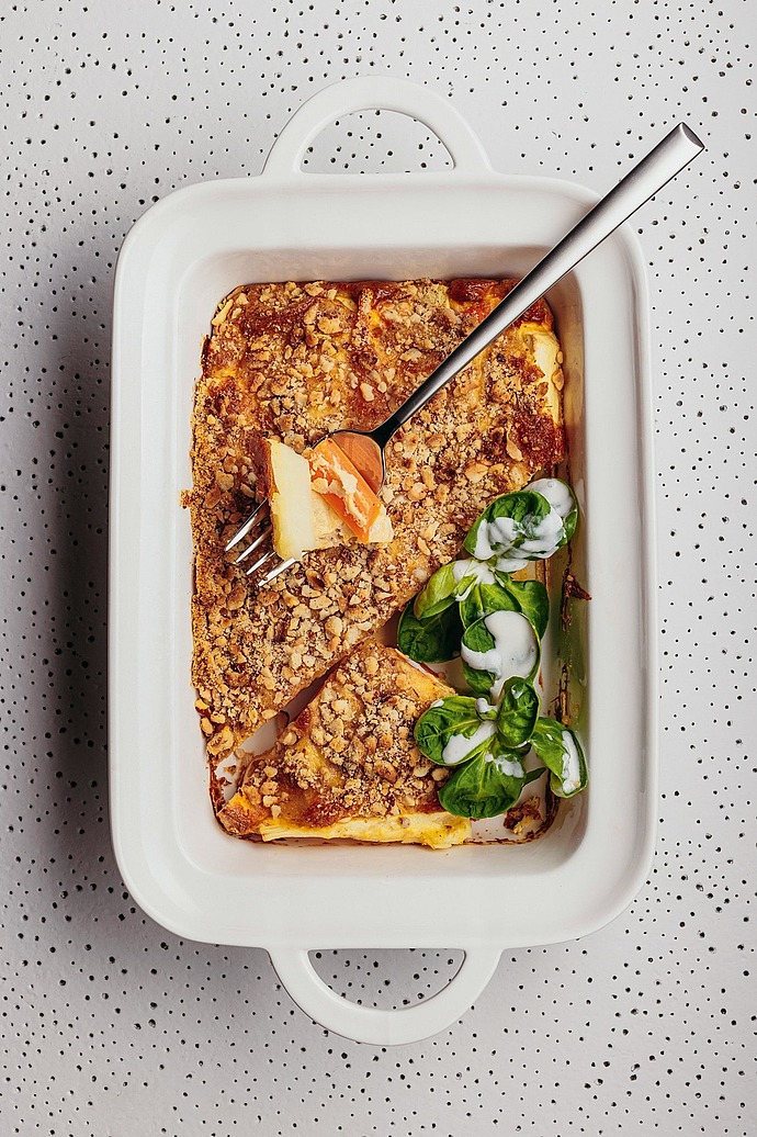 Eine weiße Auflaufform mit einem Stück Auflauf mit Krümeln und Blattspinat, der mit einer weißen Soße beträufelt ist; auf der Form liegt eine Gabel, die ein Stück des Auflaufs hält. Die Schale steht auf einer gesprenkelten Fläche.