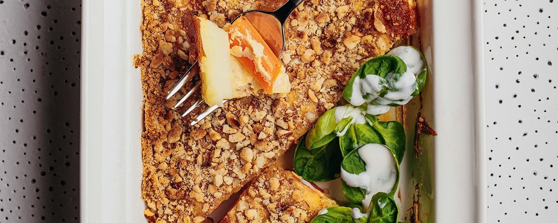 Eine weiße Auflaufform mit einem Stück Auflauf mit Krümeln und Blattspinat, der mit einer weißen Soße beträufelt ist; auf der Form liegt eine Gabel, die ein Stück des Auflaufs hält. Die Schale steht auf einer gesprenkelten Fläche.