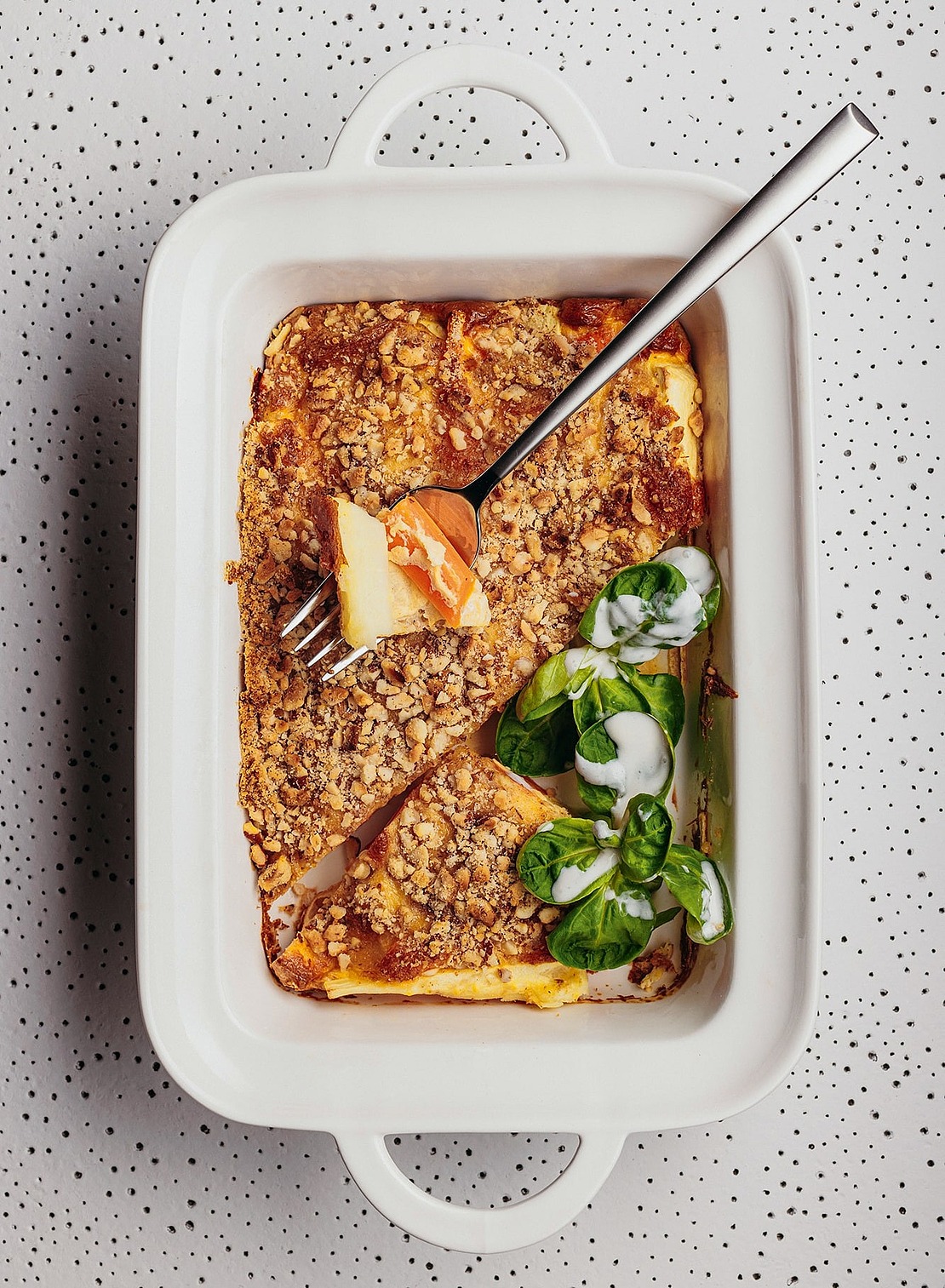 Eine weiße Auflaufform mit einem Stück Auflauf mit Krümeln und Blattspinat, der mit einer weißen Soße beträufelt ist; auf der Form liegt eine Gabel, die ein Stück des Auflaufs hält. Die Schale steht auf einer gesprenkelten Fläche.