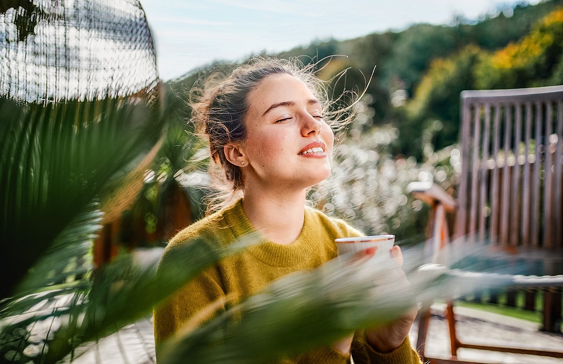 Eine Frau sitzt im Freien mit geschlossenen Augen, lächelt sanft und hält eine Tasse in der Hand. Das Sonnenlicht beleuchtet ihr Gesicht, und das grüne Laub im Vorder- und Hintergrund schafft eine friedliche und entspannte Atmosphäre.