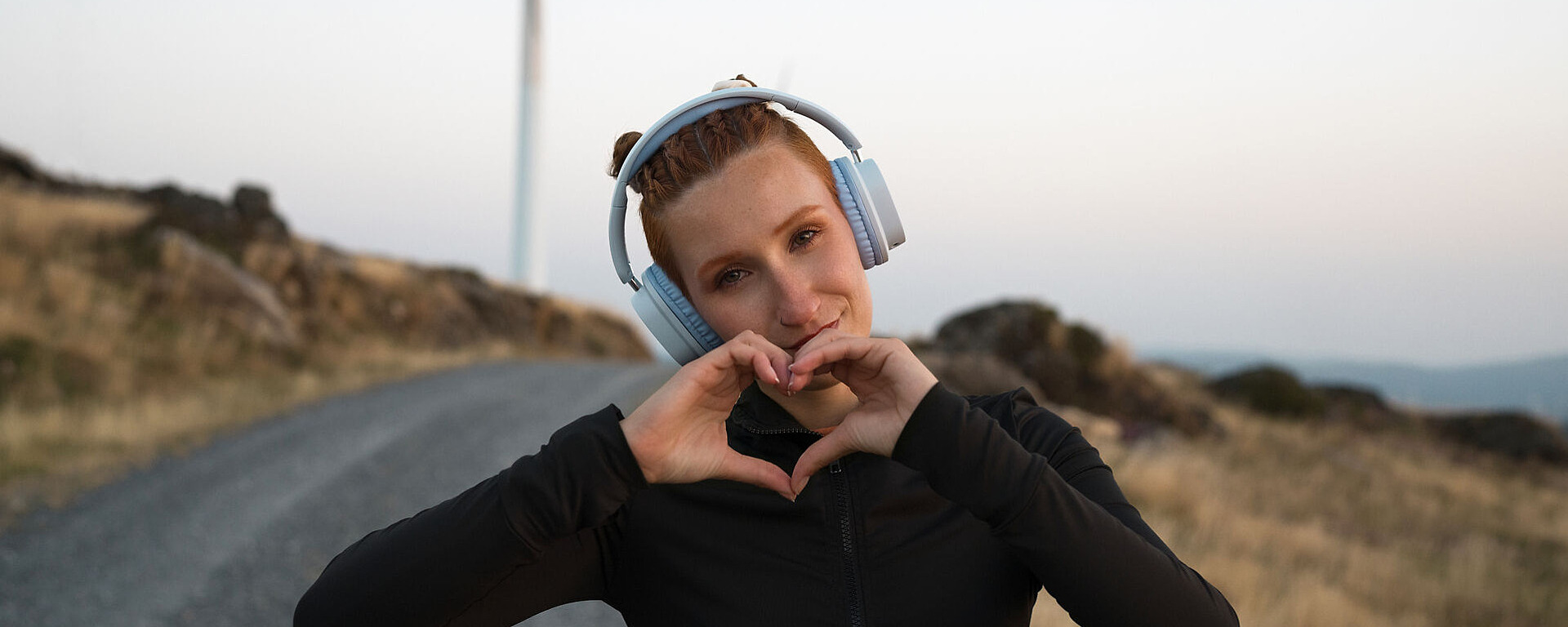Eine Frau mit Kopfhörern und schwarzem Oberteil steht auf einem Kiesweg im Freien und bildet mit ihren Händen eine Herzform. Der Hintergrund ist eine grasbewachsene, felsige Landschaft unter einem klaren Himmel.