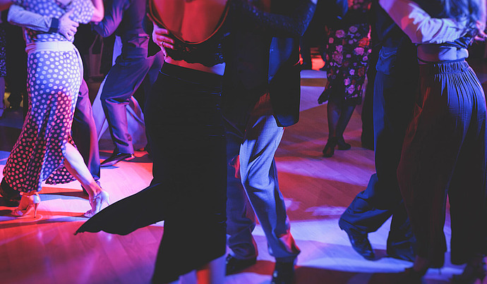 Paare tanzen eng aneinander gedrängt in einem schwach beleuchteten Raum mit bunten Lichtern, die violette, rosa und blaue Farbtöne erzeugen. Sie tragen elegante Abendgarderobe, die ihre Bewegung und Anmut auf dem polierten Holzboden betont.