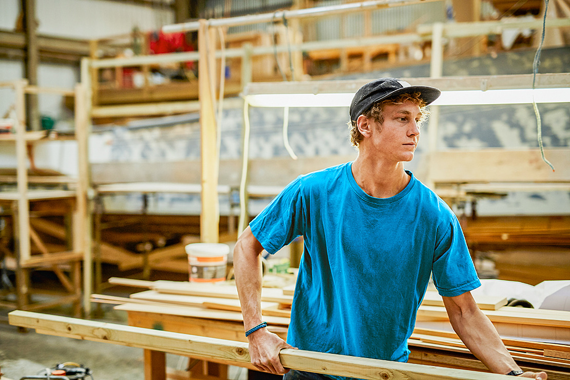 Ein junger Mann in blauem Hemd und Mütze arbeitet in einer Schreinerei und hält einen Holzbalken in der Hand. Er ist von Werkzeugen und verschiedenen Holzstücken umgeben. Im Hintergrund sind Regale und eine Werkbank unter heller Beleuchtung zu sehen.