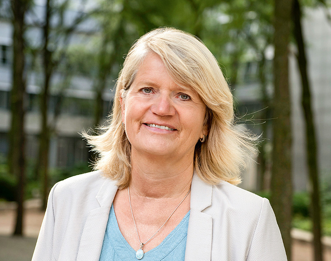 Eine Frau mit blondem Haar, die ein hellblaues Oberteil und einen hellen Blazer trägt, steht im Freien und lächelt, während im Hintergrund Bäume und moderne Gebäude zu sehen sind.