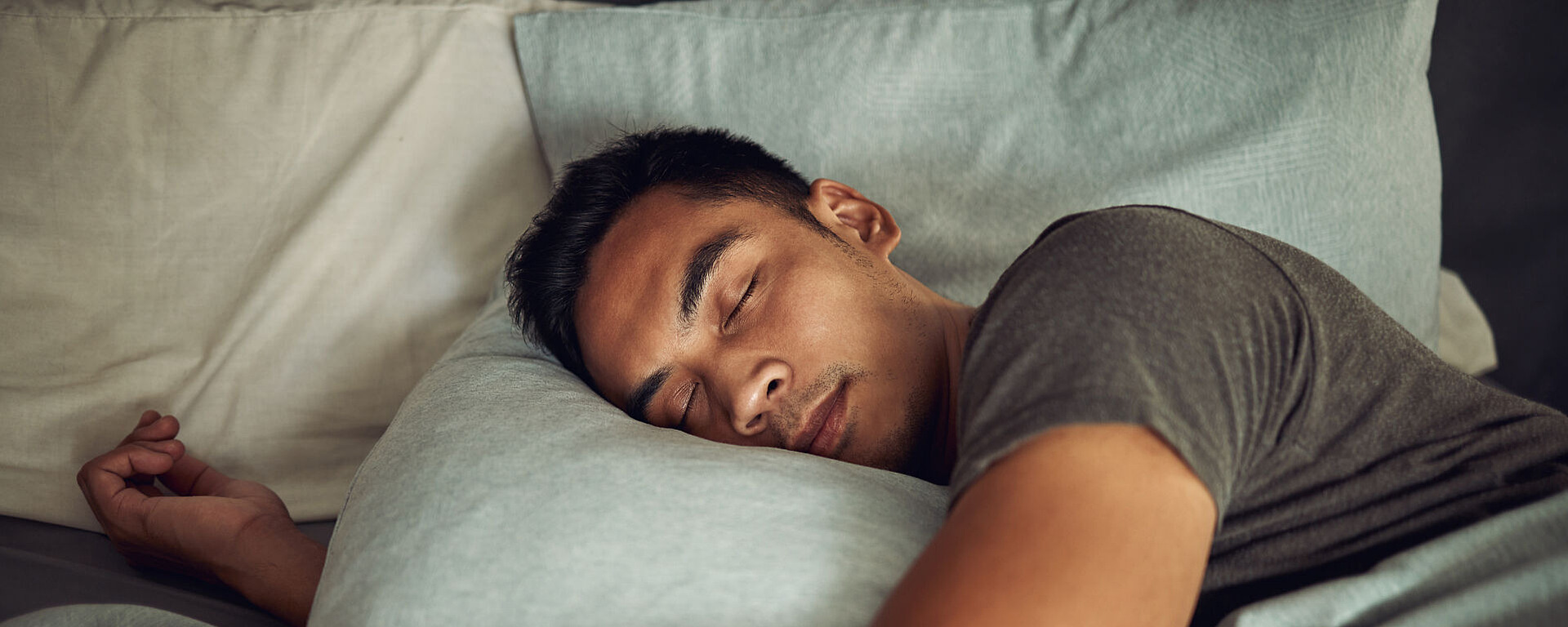 Ein Mann schläft seitlich im Bett, sein Kopf ruht auf einem hellblauen Kissen. Er trägt ein graues T-Shirt und wirkt friedlich und entspannt.