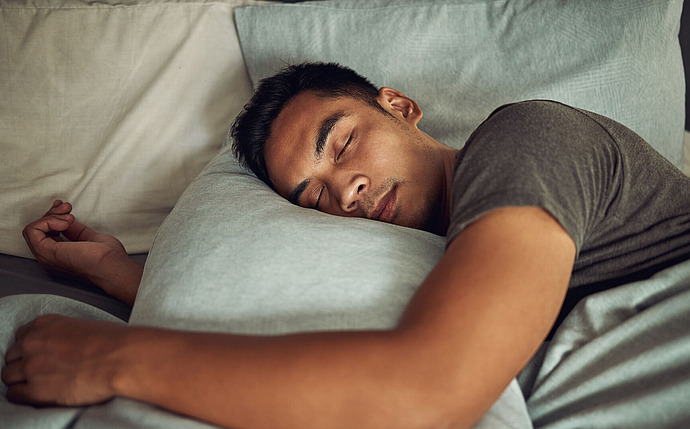 Ein Mann schläft seitlich im Bett, sein Kopf ruht auf einem hellblauen Kissen. Er trägt ein graues T-Shirt und wirkt friedlich und entspannt.