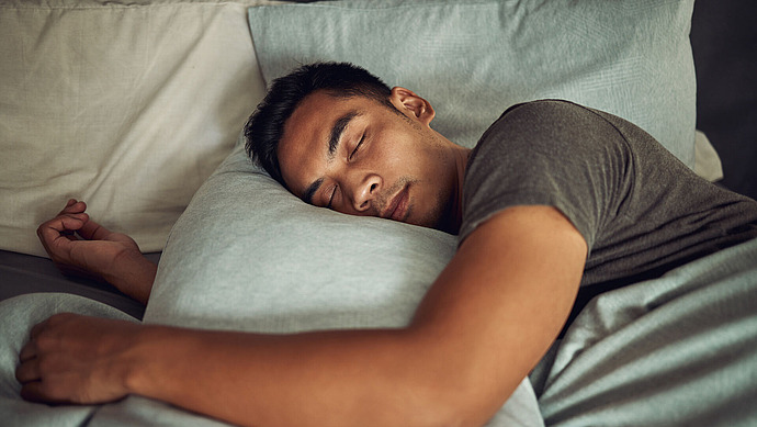 Ein Mann schläft seitlich im Bett, sein Kopf ruht auf einem hellblauen Kissen. Er trägt ein graues T-Shirt und wirkt friedlich und entspannt.