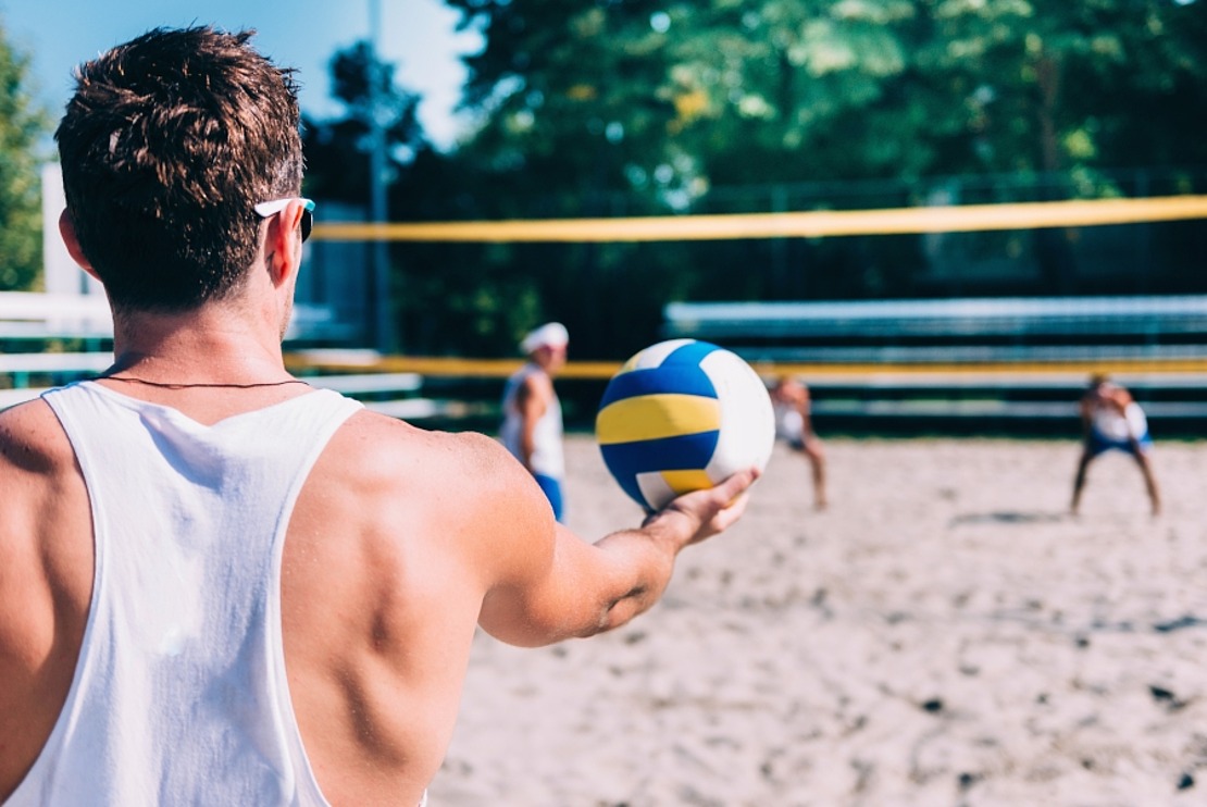 Ein Mann in einem weißen Tank-Top bereitet sich darauf vor, einen Volleyball auf einem sandigen Strandplatz aufzuschlagen, während drei Gegner auf der anderen Seite des Netzes unter einem sonnigen Himmel bereitstehen.