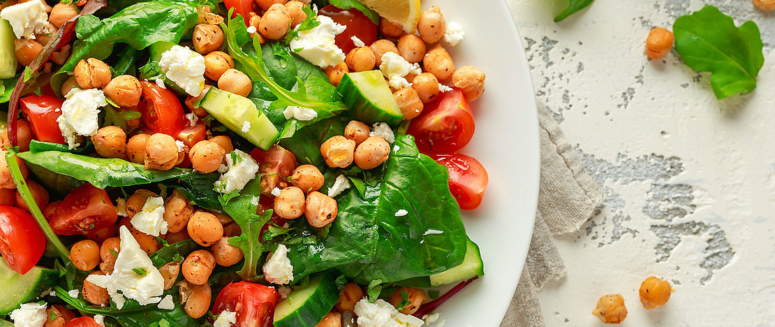 Ein frischer Salat mit Spinat, Kirschtomaten, Gurke, Kichererbsen, zerbröckeltem Feta und einer Zitronenscheibe auf einem weißen Teller, platziert auf einer leicht strukturierten Oberfläche. Daneben liegen einige Kichererbsen und Blattgemüse.