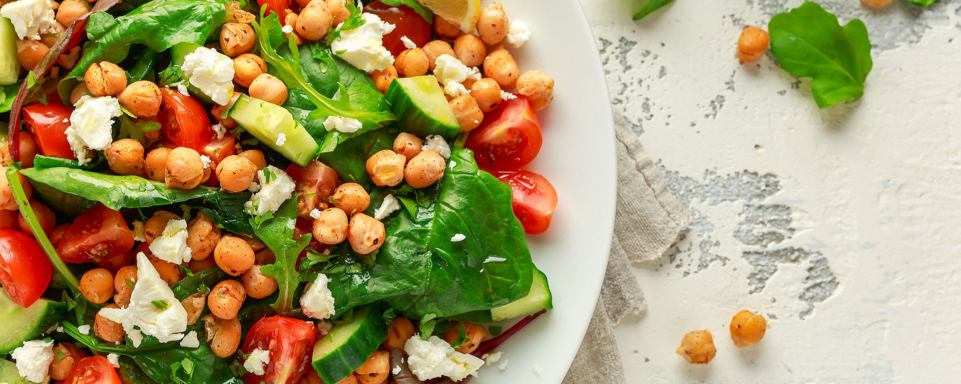 Ein frischer Salat mit Spinat, Kirschtomaten, Gurke, Kichererbsen, zerbröckeltem Feta und einer Zitronenscheibe auf einem weißen Teller, platziert auf einer leicht strukturierten Oberfläche. Daneben liegen einige Kichererbsen und Blattgemüse.