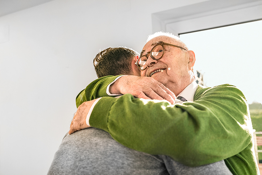 Ein älterer Mann mit Brille und grünem Pullover lächelt fröhlich, während er in einem sonnendurchfluteten Raum eine andere Person umarmt.