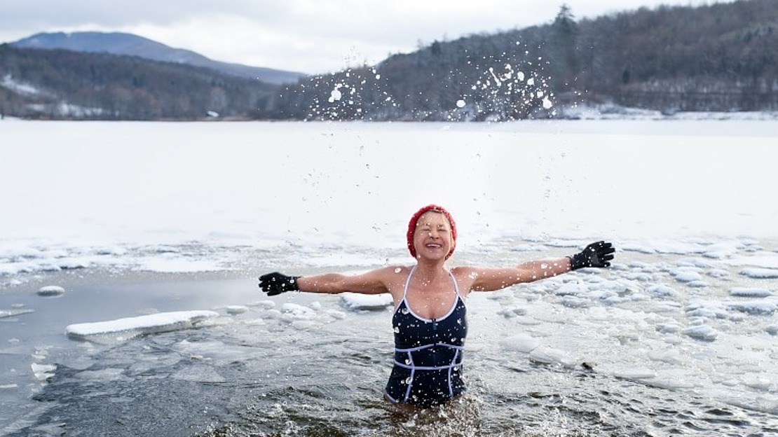 Eine Person im Badeanzug und mit roter Mütze steht in einem eisigen See, hebt die Arme und spritzt mit dem Wasser herum. Schnee und Eis bedecken den See und die umliegende Landschaft, mit Hügeln und Bäumen im Hintergrund.