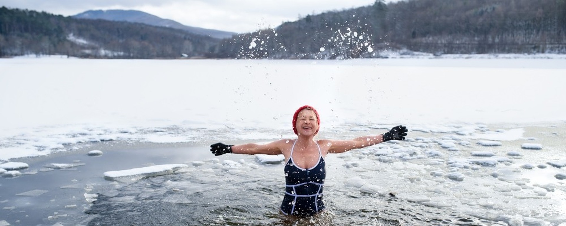 Eine Person im Badeanzug und mit roter Mütze steht in einem eisigen See, hebt die Arme und spritzt mit dem Wasser herum. Schnee und Eis bedecken den See und die umliegende Landschaft, mit Hügeln und Bäumen im Hintergrund.
