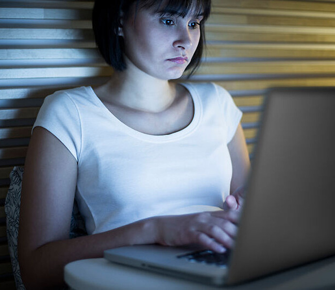 Eine junge Frau sitzt in einem Haus, trägt ein weißes T-Shirt und schaut konzentriert auf einen Laptop-Bildschirm bei schwacher Beleuchtung, mit Holzlatten im Hintergrund.