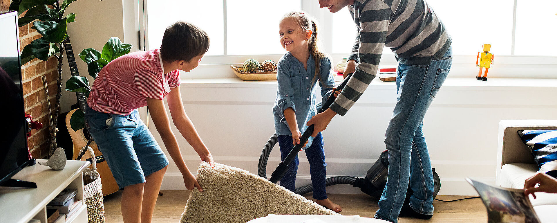Ein Vater und zwei Kinder saugen und räumen ein helles Wohnzimmer auf. Vater und Tochter bedienen den Staubsauger, während der Sohn einen kleinen Teppich hochhebt. Der Raum ist mit Pflanzen und Spielzeug dekoriert, Sonnenlicht fällt durch das Fenster.