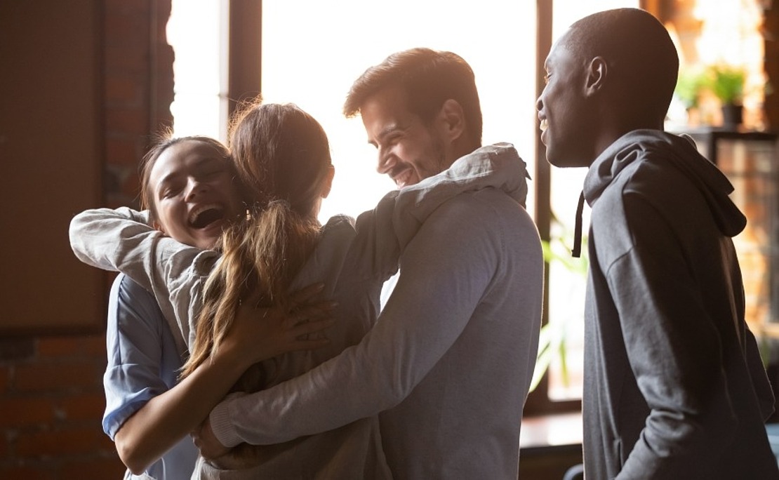 Vier junge Erwachsene stehen in einem hellen Raum, lächeln, umarmen sich herzlich und scheinen glücklich und zufrieden zu sein.