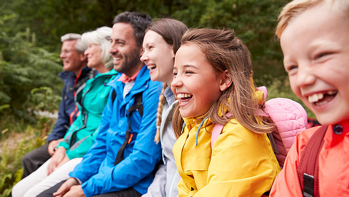 Eine generationsübergreifende Gruppe, darunter Kinder und Erwachsene, sitzt in bunten Jacken im Freien, lächelt und lacht zusammen, während im Hintergrund Grünflächen zu sehen sind.