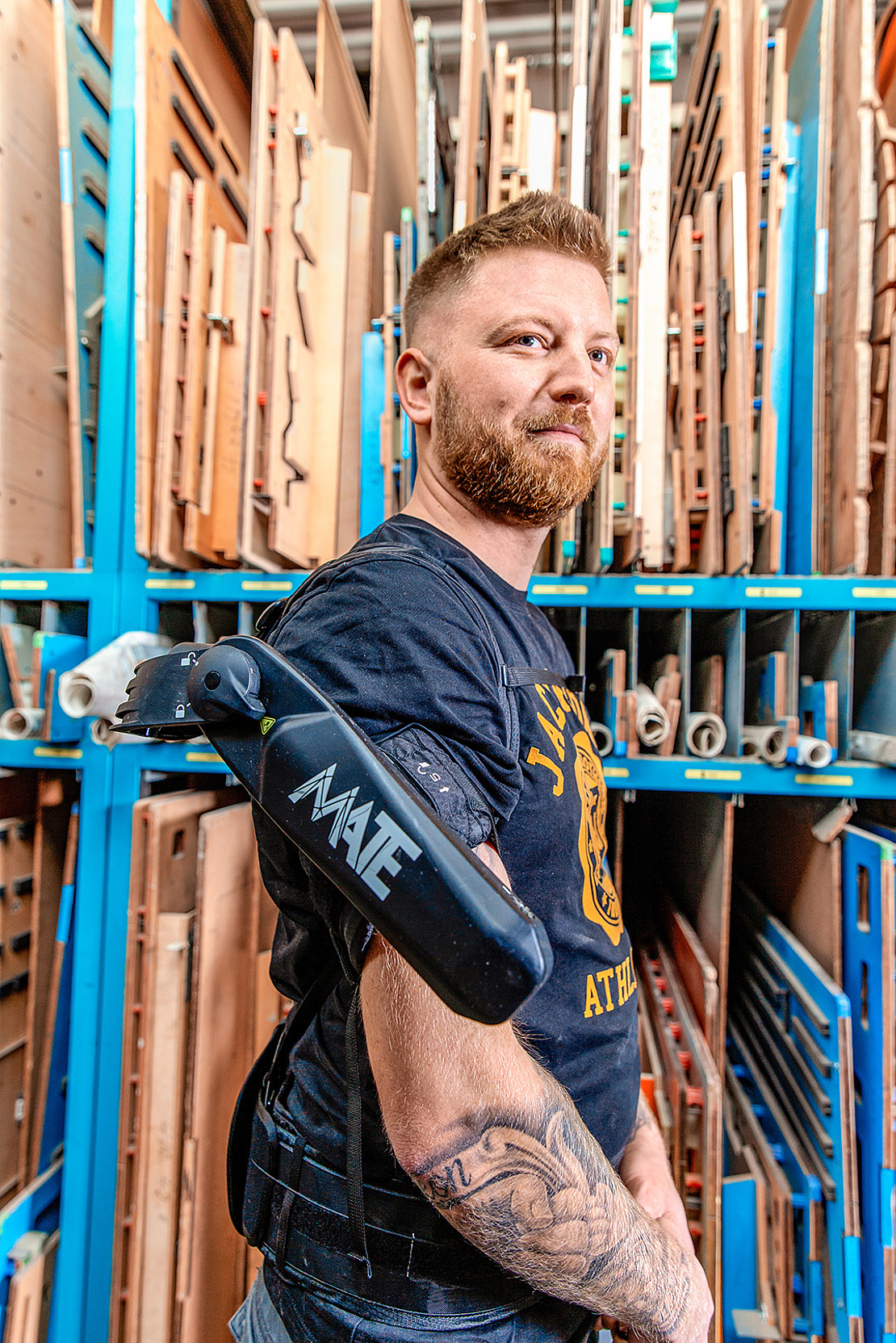 Ein bärtiger Mann steht vor Regalen mit Holzbrettern und trägt einen schwarzen Exoskelettarm mit der Aufschrift "MATE". Er hat Tätowierungen auf seinem rechten Arm und blickt selbstbewusst in die Kamera.