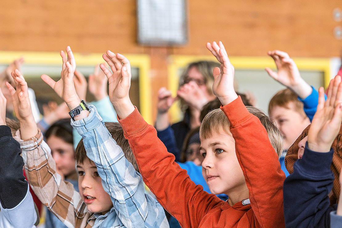 Eine Gruppe von Kindern in einem Klassenzimmer hebt enthusiastisch die Hände, einige lächeln, während sie nach vorne schauen. Der Hintergrund ist unscharf und hebt die Gesichter und ausgestreckten Arme der Kinder hervor.