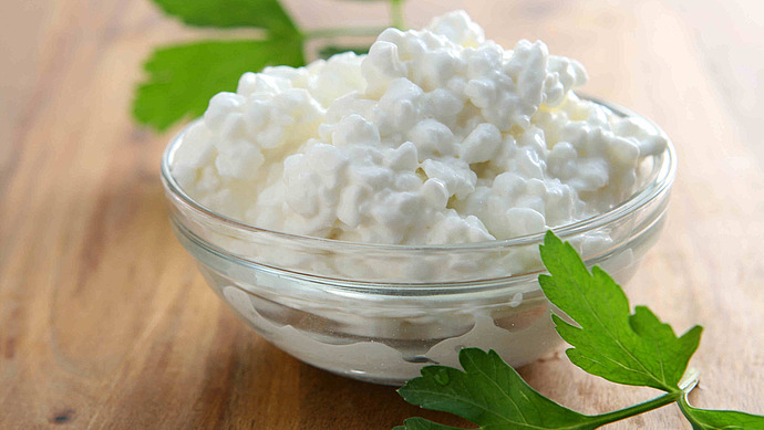 Eine mit Hüttenkäse gefüllte Glasschale steht auf einer Holzplatte. Frische grüne Petersilienblätter werden zur Dekoration um die Schale herum und teilweise darunter gelegt.