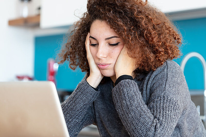 Eine junge Frau mit lockigem Haar sitzt an einem Tisch, hat den Kopf in die Hände gestützt und schaut mit gelangweiltem oder müdem Gesichtsausdruck auf einen Laptop-Bildschirm. Sie trägt einen grauen Pullover und sitzt in einer Küche.