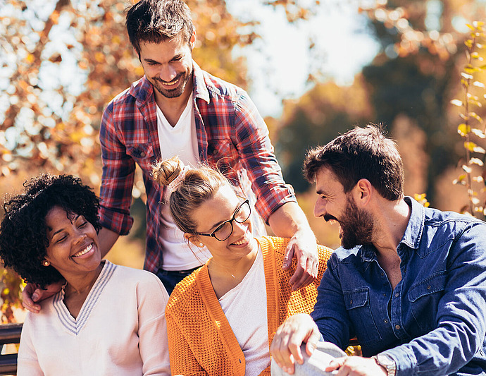 Vier Freunde sitzen und stehen im Herbst zusammen auf einer Bank im Freien, lächeln und lachen zwischen goldenem Laub. Zwei Frauen sitzen, zwei Männer stehen und beugen sich vor. Alle wirken glücklich und genießen die Gesellschaft des anderen.