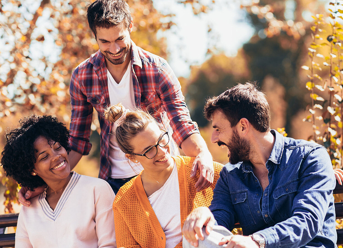 Vier Freunde sitzen und stehen im Herbst zusammen auf einer Bank im Freien, lächeln und lachen zwischen goldenem Laub. Zwei Frauen sitzen, zwei Männer stehen und beugen sich vor. Alle wirken glücklich und genießen die Gesellschaft des anderen.