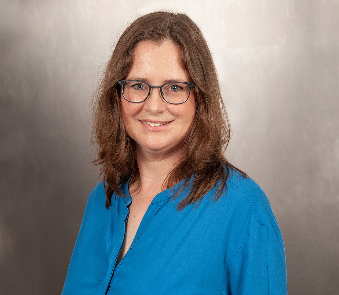 Eine Frau mit schulterlangem, braunem Haar und Brille, die eine hellblaue Bluse trägt, steht lächelnd vor einem neutralen, sanft beleuchteten Hintergrund.