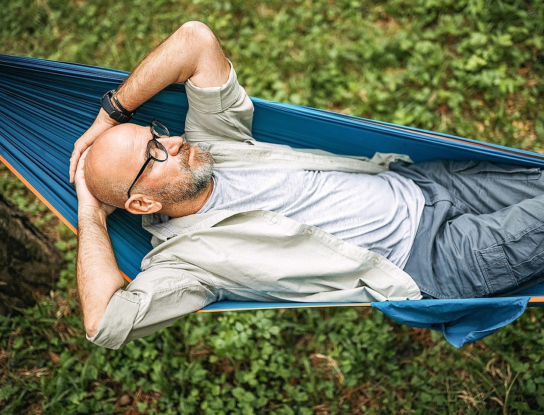 Ein glatzköpfiger Mann mit Brille entspannt sich in einer blauen Hängematte im Freien, auf dem Rücken liegend, die Hände hinter dem Kopf, umgeben von grünem Gras und Bäumen.