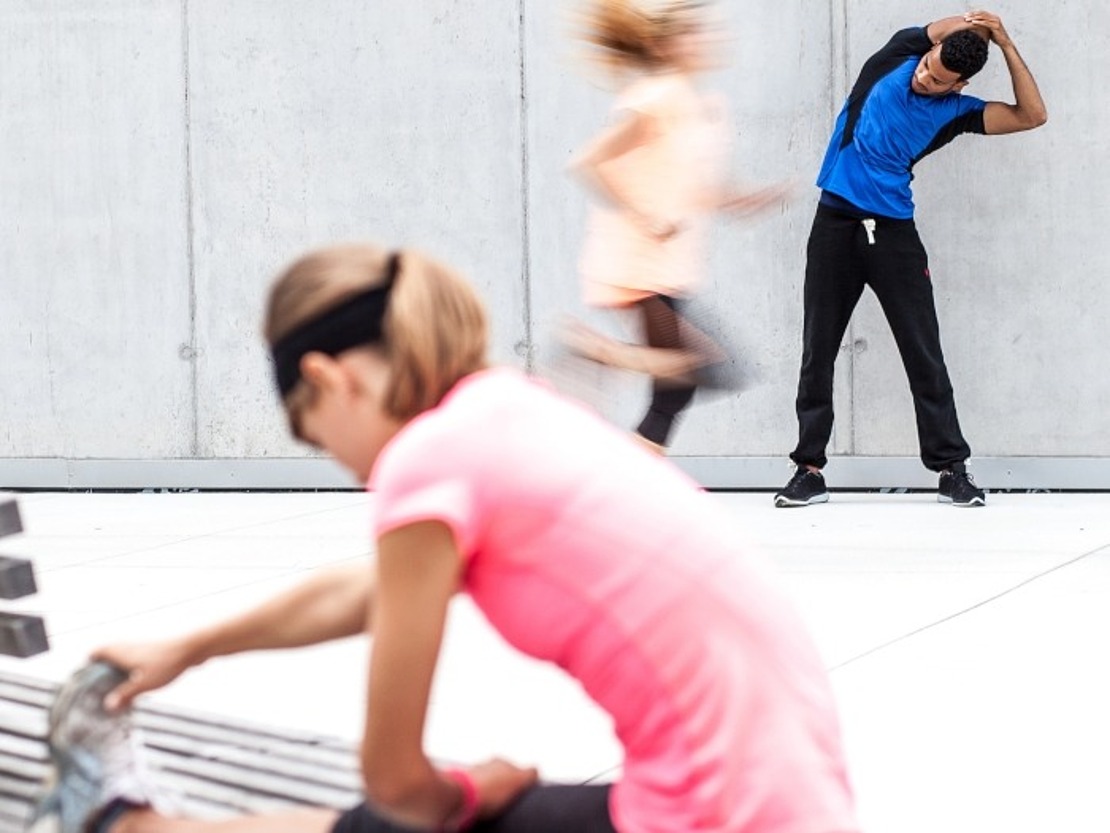 Eine Frau in einem rosafarbenen Hemd streckt sich auf einer Bank im Vordergrund, während ein Mann in einem blauen Hemd sich gegen eine graue Wand streckt und im Hintergrund zwei unscharfe Läufer vorbeiziehen.