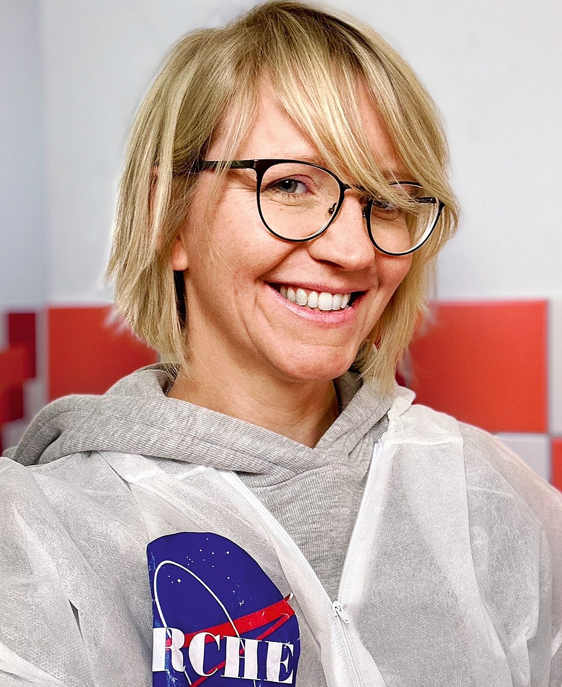 Eine lächelnde Frau mit kurzen blonden Haaren und Brille trägt einen weißen Laborkittel mit einem wissenschaftlichen Logo und einen grauen Kapuzenpulli und steht vor einer rot-weiß gekachelten Wand.