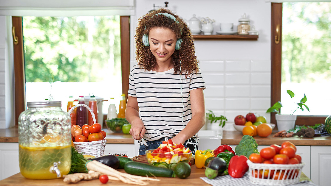 Eine Frau mit Kopfhörern lächelt, während sie in einer hellen Küche Gemüse schneidet. Die Arbeitsplatte ist mit verschiedenen frischen Produkten gefüllt, darunter Tomaten, Paprika und Gurken, und daneben steht ein großes Glas mit Limonade.