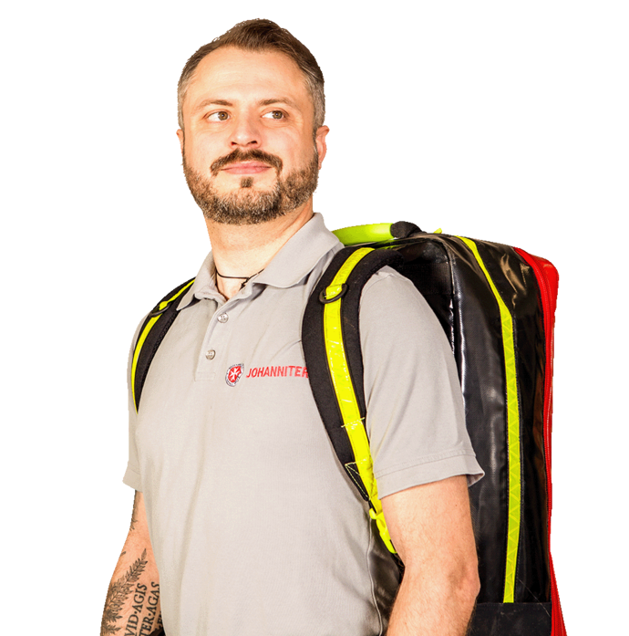 Ein Mann, Notfallsanitäter Maximilian Class aus Villingen-Schwenningen, in einem grauen Poloshirt und einem gelb-schwarzen Rettungsrucksach steht mit leicht nach links geneigtem Gesicht vor einem schlichten weißen Hintergrund und blickt zur Seite.