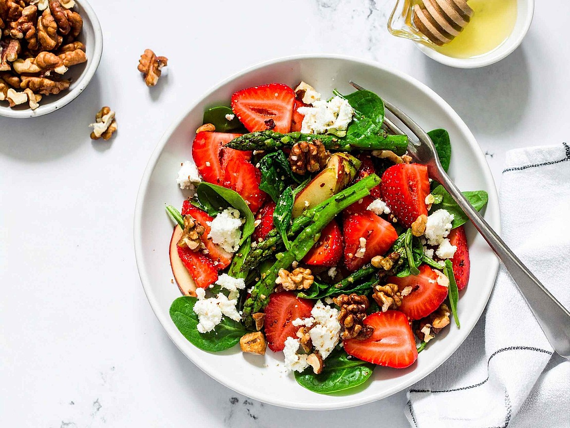 Eine Schüssel Spinatsalat mit geschnittenen Erdbeeren, geröstetem Spargel, zerbröckeltem Feta und Walnüssen, daneben eine Gabel. Daneben stehen eine kleine Schale mit Walnüssen und ein Glas Honig auf einer Marmorplatte.