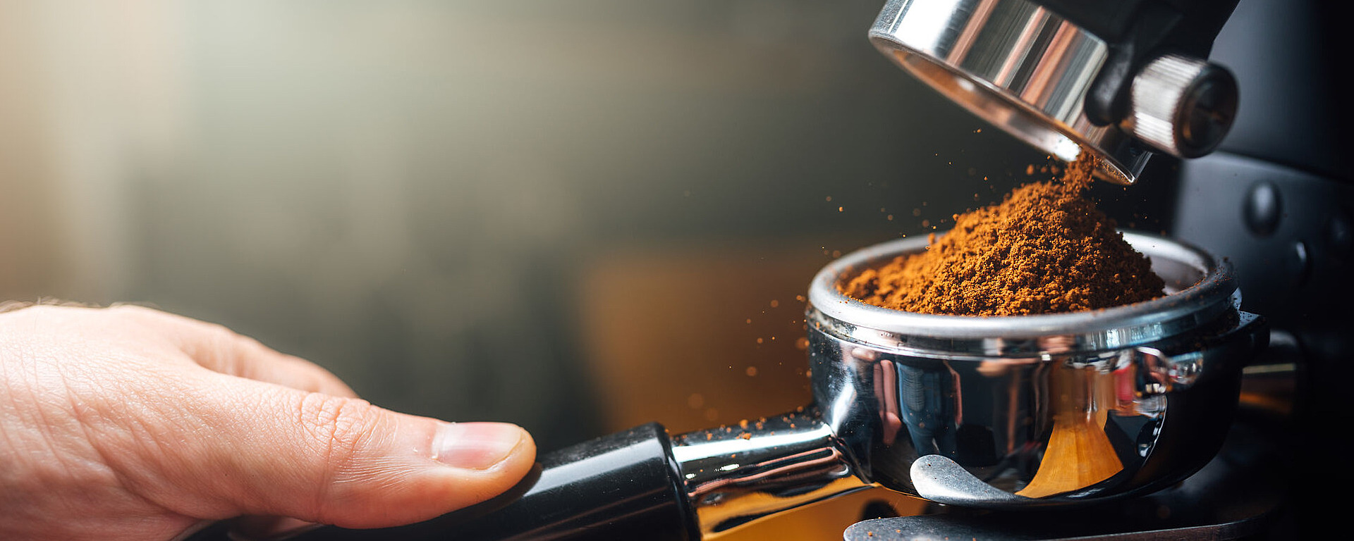 Eine Nahaufnahme einer Hand, die einen Siebträger unter einer Mühle hält, während frisch gemahlener Kaffee in den Siebträger fällt und zum Aufbrühen in einer Espressomaschine bereit ist.