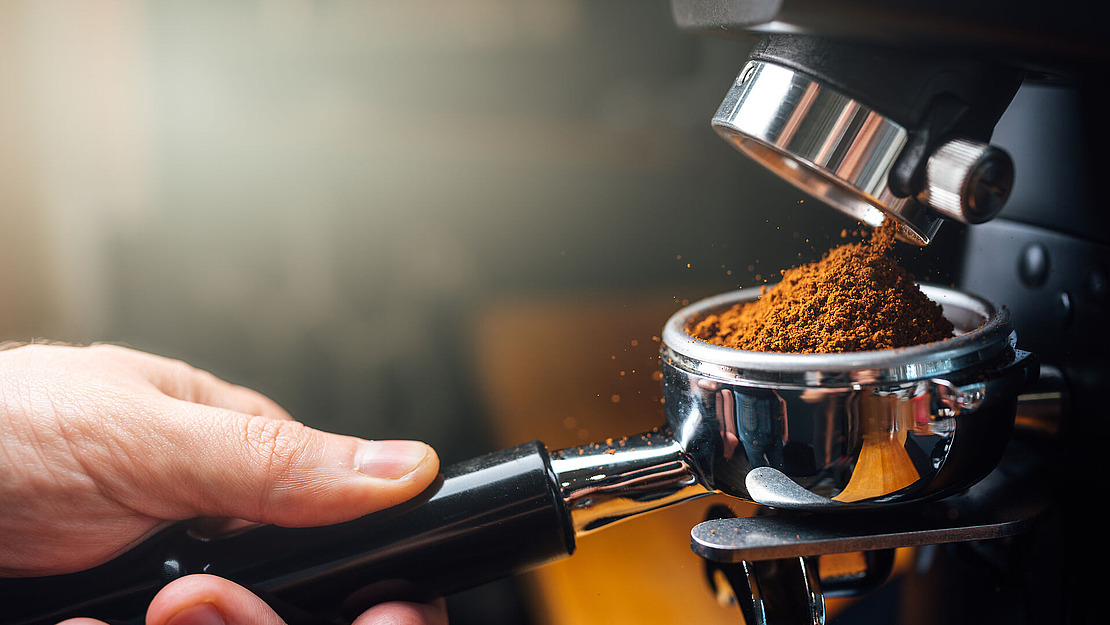 Eine Nahaufnahme einer Hand, die einen Siebträger unter einer Mühle hält, während frisch gemahlener Kaffee in den Siebträger fällt und zum Aufbrühen in einer Espressomaschine bereit ist.