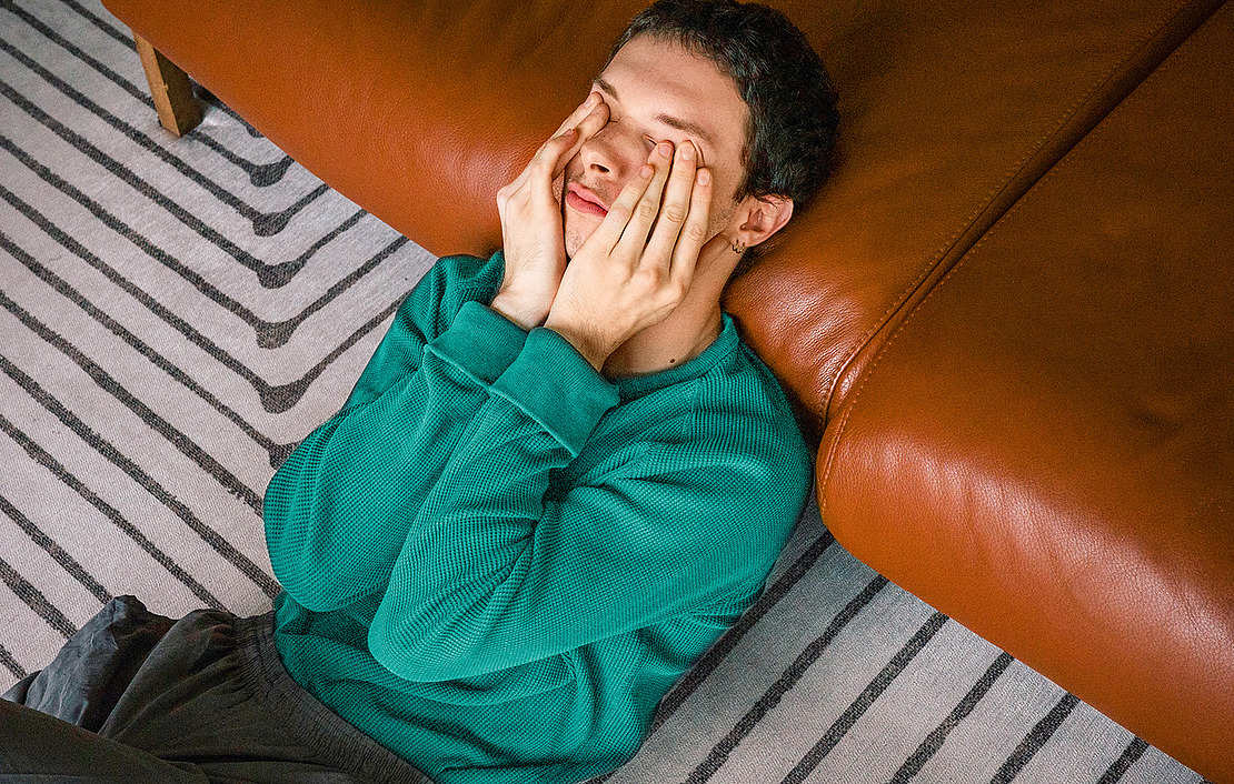 Eine Person in einem grünen Pullover sitzt auf einem gestreiften Teppich, an ein braunes Ledersofa gelehnt, mit geschlossenen Augen und Händen, die das Gesicht bedecken, und scheint gestresst oder müde zu sein.