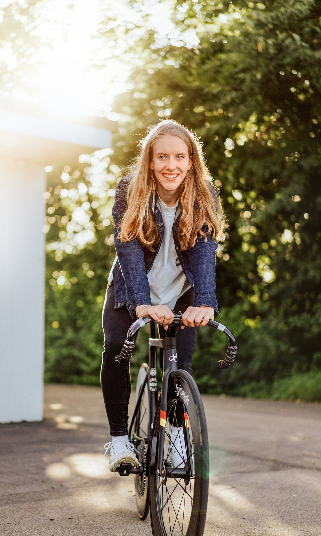 Eine junge Frau mit langen blonden Haaren fährt an einem sonnigen Tag auf einem schwarzen Fahrrad und lächelt in die Kamera. Sie trägt eine blaue Jacke, ein helles Hemd und eine Jeans. Grüne Bäume und Sonnenlicht füllen den Hintergrund.