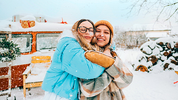 Zwei Freundinnen lächeln und umarmen sich in einer verschneiten Umgebung. Eine trägt eine blaue Pufferjacke und eine Brille, die andere Person einen karierten Mantel und eine Strickmütze. Im Hintergrund sind ein Oldtimer-Wohnmobil und ein Stapel Brennholz zu sehen, um sie herum fällt sanft Schnee.