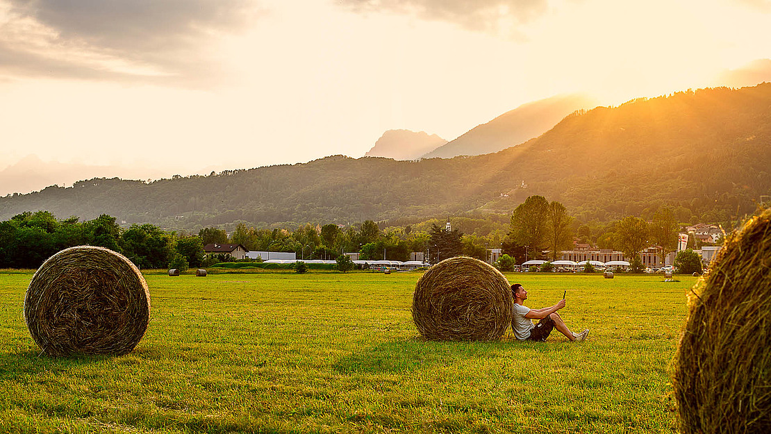 Eine Person sitzt im Gras, an einen großen Heuballen gelehnt, und macht ein Selfie in einem sonnenbeschienenen Feld mit weiteren Heuballen, Bergen, Bäumen und Gebäuden im Hintergrund während des Sonnenuntergangs.